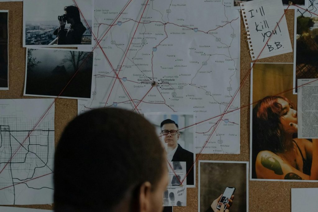 Analyst studying a protective intelligence case board with photographs, maps, and connecting lines linking persons of interest to locations